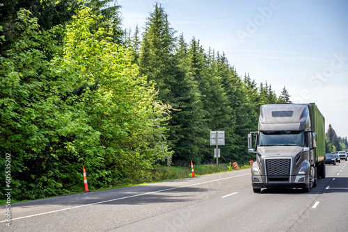 Dark gray big rig semi truck tractor transporting loaded container on semi trailer driving on the straight highway road with trees on the side