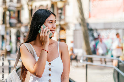 Latin American female traveler with backpack on shoulder looking away with smile and answering phone call on blurred background of sunlit street on weekend in Barcelona, Spain © dzmatph
