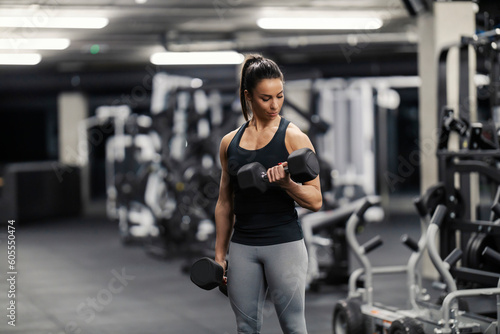 A sportswoman in shape is doing exercises for biceps while flexing muscles in a gym.