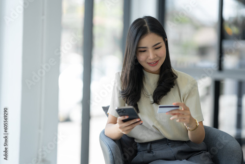 Confident young asian woman using mobile phone to open application using credit card for online shopping while sitting on sofa chair at office.