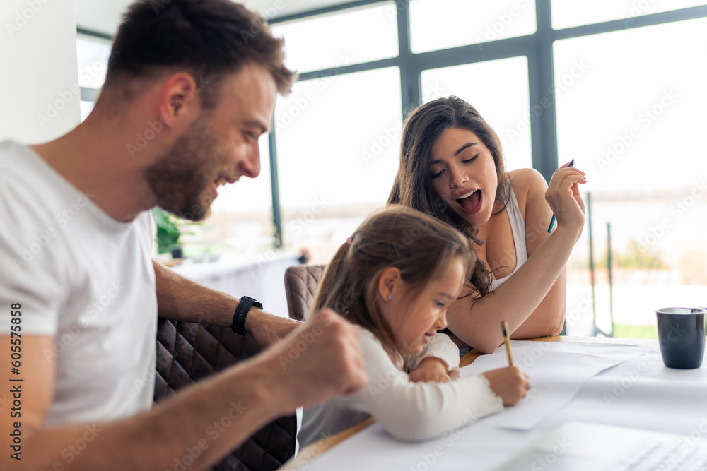 Young parents teach their daughter to write, helping her with her homework, untidy desk and laptop. Concept happy healthy family Young parents teach their daughter to write, helping her with her homework, untidy desk and laptop. Concept happy healthy family