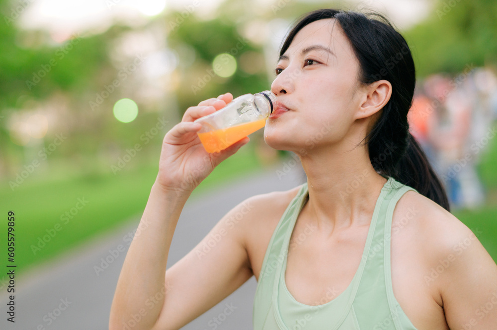 Female jogger. Fit young Asian woman with green sportswear drinking organic orange juice after running and enjoying a healthy outdoor. Fitness runner girl in public park. Wellness being concept