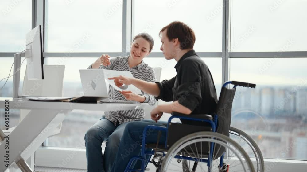 Young male office worker in a wheelchair talking to female colleague while showing her ideas using laptop in co-working space. Disability and handicap concept. Office modern people at work. Young male office worker in a wheelchair talking to female colleague while showing her ideas using laptop in co-working space. Disability and handicap concept. Office modern people at work.