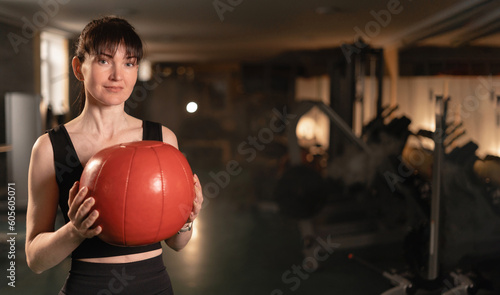 Portrait of mid adult woman looking at camera while holding heavy medicine ball over gym background. Strong woman with heavy ball after cross training workout.
