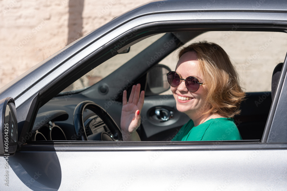 A smiling girl in sunglasses sitting in a car and waving her hand, a welcoming gesture. The concept of traveling by car.