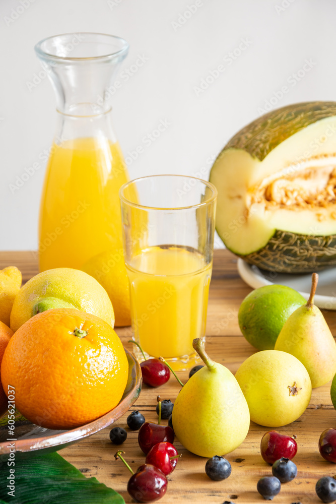 Close-up of bottle and glass with orange juice with melon, oranges, lemons, cherries, pears and blueberries on table, white background, vertical, with copy space Close-up of bottle and glass with orange juice with melon, oranges, lemons, cherries, pears and blueberries on table, white background, vertical, with copy space