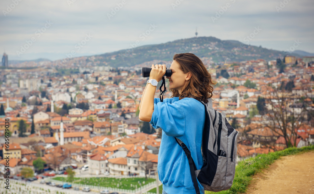 A young adult smiling female traveler looks through binoculars at a European city with orange roofs in the mountains. Travel to the Balkans, Sarajevo, Bosnia A young adult smiling female traveler looks through binoculars at a European city with orange roofs in the mountains. Travel to the Balkans, Sarajevo, Bosnia