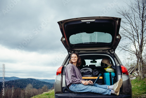 Young pretty smiling woman traveler in casual wear sitting in car trunk using laptop while traveling in mountains