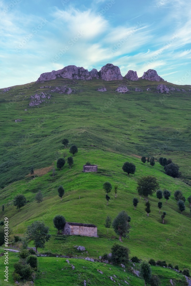 Fototapeta premium Small stone cottage in lush green countryside with hills under blue cloudy sky in Cantabria, Spain