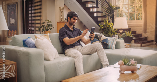 Cheerful Young Indian Man Comfortably Using His Smartphone At Home, Hansdome, Smiling and Positive. He's Scrolling Through Social Media Or Engaging In Virtual Remote Work.