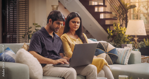Adorable Young Indian Couple Using Laptop, Sitting on Home Sofa. Exploring Online Content, Discussing Trend Ideas and Making Purchase Decisions Together. Online Shopping Concept.  © Kitreel
