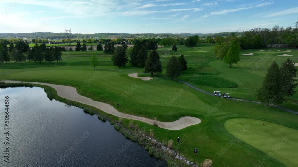 Aerial drone shot of green golf course and pond on a sunny day. Blue sky above rural farmland and manicured golf course. Large sand traps and water hazard featured. Golfers enjoy a day on the green. Aerial drone shot of green golf course and pond on a sunny day. Blue sky above rural farmland and manicured golf course. Large sand traps and water hazard featured. Golfers enjoy a day on the green.