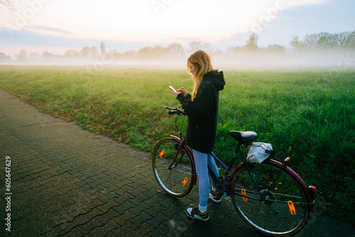 Young attractive caucasian woman using mobile phone, ride on bicycle in the park in sunset with fog .