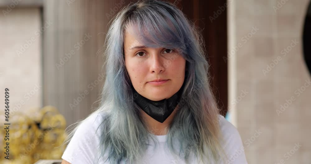 Young woman with colored hair wear black mask, portrait shot with blurred background, she look straight in camera. Then girl pull mask down to open nose and mouth and make sigh Young woman with colored hair wear black mask, portrait shot with blurred background, she look straight in camera. Then girl pull mask down to open nose and mouth and make sigh