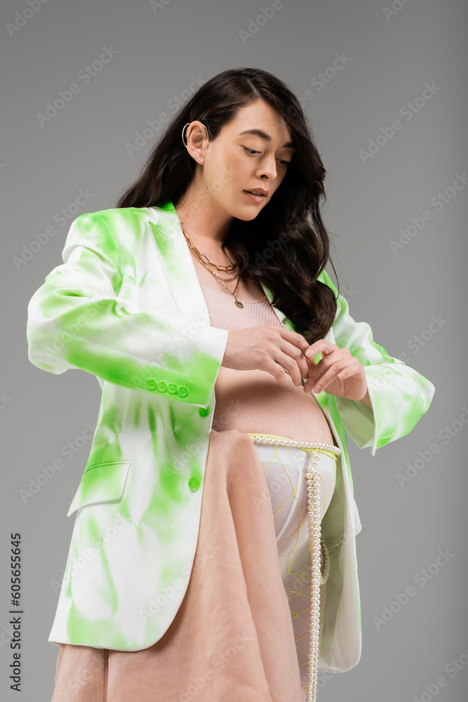 expecting mother in green and white jacket, crop top, leggings with beads belt and chiffon cloth posing with hands near tummy isolated on grey background, fashionable maternity concept expecting mother in green and white jacket, crop top, leggings with beads belt and chiffon cloth posing with hands near tummy isolated on grey background, fashionable maternity concept