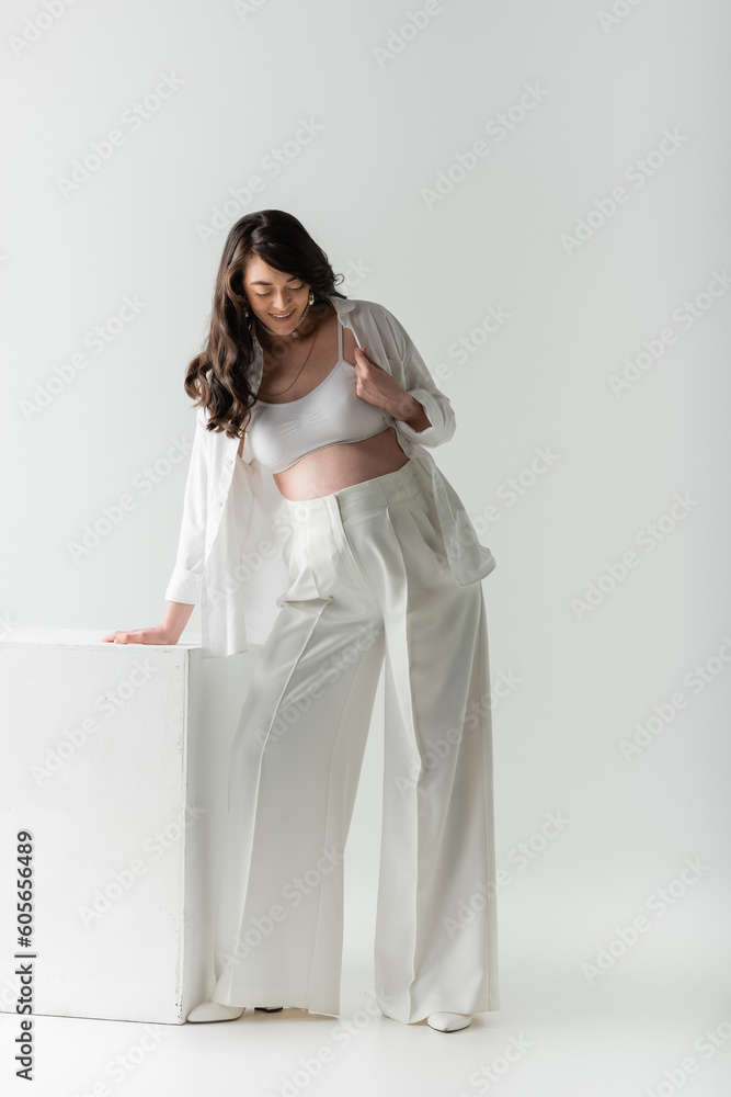 full length of pleased pregnant woman with wavy brunette hair, wearing white pants, crop top and shirt, standing near cube on grey background, maternity fashion concept full length of pleased pregnant woman with wavy brunette hair, wearing white pants, crop top and shirt, standing near cube on grey background, maternity fashion concept