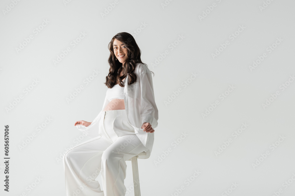 cheerful pregnant woman in white fashionable clothes such as white shirt and pants sitting on stool and smiling at camera isolated on grey background, maternity fashion concept cheerful pregnant woman in white fashionable clothes such as white shirt and pants sitting on stool and smiling at camera isolated on grey background, maternity fashion concept