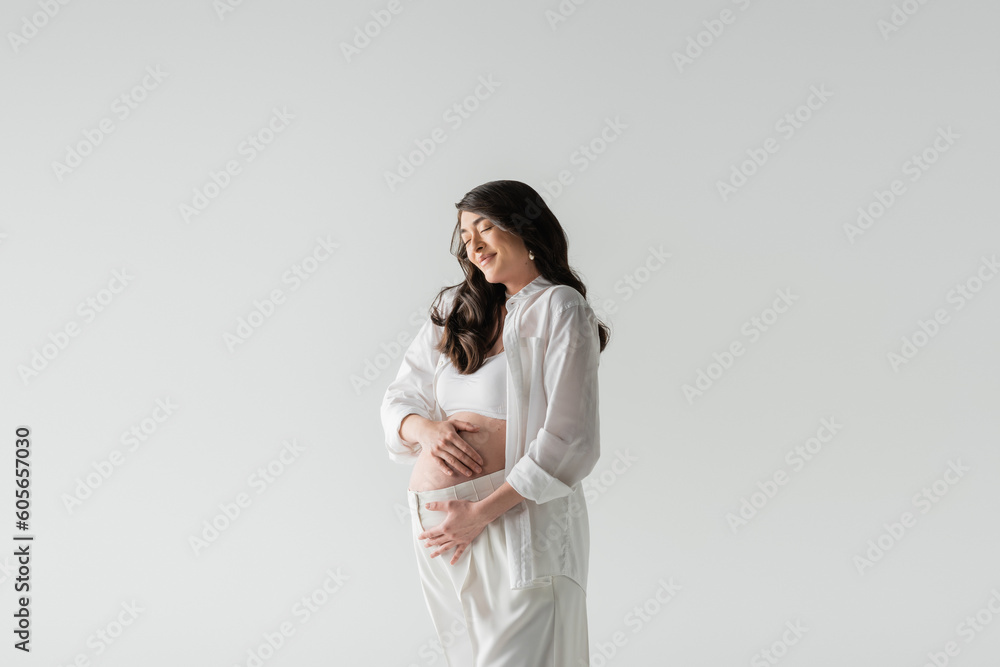 appealing and stylish pregnant model in white crop top, shirt and pants embracing tummy and smiling with closed eyes isolated on grey background, maternity fashion concept appealing and stylish pregnant model in white crop top, shirt and pants embracing tummy and smiling with closed eyes isolated on grey background, maternity fashion concept