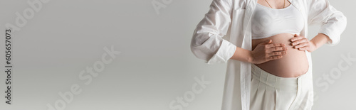 cropped view of trendy pregnant woman touching belly while posing in white crop top, shirt and pants isolated on grey background, maternity fashion concept, banner © LIGHTFIELD STUDIOS