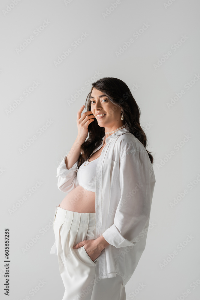 charming pregnant woman with wavy brunette hair, in white shirt, crop top and pants, smiling and looking at camera isolated on grey background, maternity fashion concept charming pregnant woman with wavy brunette hair, in white shirt, crop top and pants, smiling and looking at camera isolated on grey background, maternity fashion concept