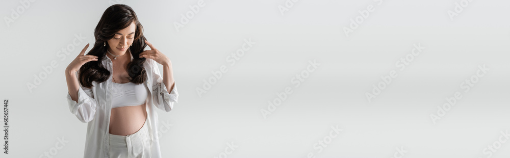 stylish pregnant woman in white trendy shirt, crop top and pants touching wavy brunette hair and posing isolated on grey background, fashionable maternity concept, banner stylish pregnant woman in white trendy shirt, crop top and pants touching wavy brunette hair and posing isolated on grey background, fashionable maternity concept, banner