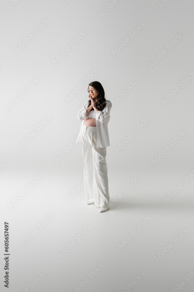 full length of trendy future mother in white fashionable pants and shirt standing with closed eyes and smiling on grey background, maternity style concept, pregnancy full length of trendy future mother in white fashionable pants and shirt standing with closed eyes and smiling on grey background, maternity style concept, pregnancy