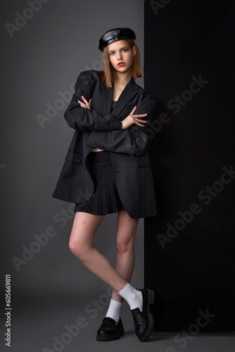 a beautiful girl with a beautiful figure in a short skirt, a hat and a large men's jacket pose standing in the studio on a dark background