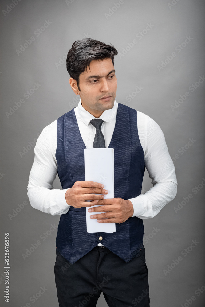 Indian young business man giving presentation on a topic holding folder in hand in formal wear wearing white shirt, black tie, black coat against grey background. Male model. Indian young business man giving presentation on a topic holding folder in hand in formal wear wearing white shirt, black tie, black coat against grey background. Male model.