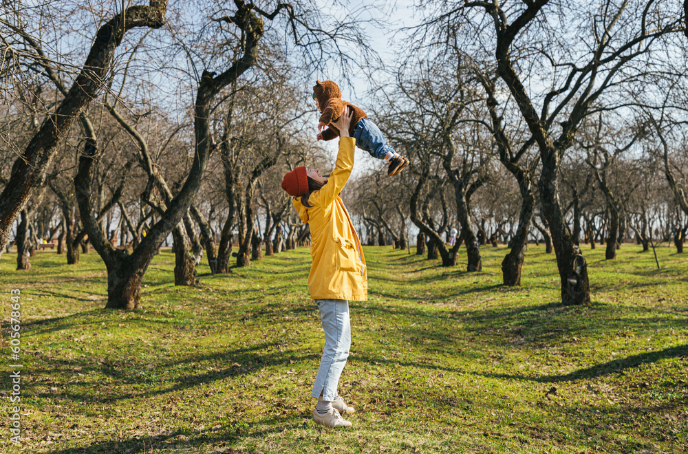 a young mother throws her child into the air against the background of an alley of trees without leaves and green grass a young mother throws her child into the air against the background of an alley of trees without leaves and green grass