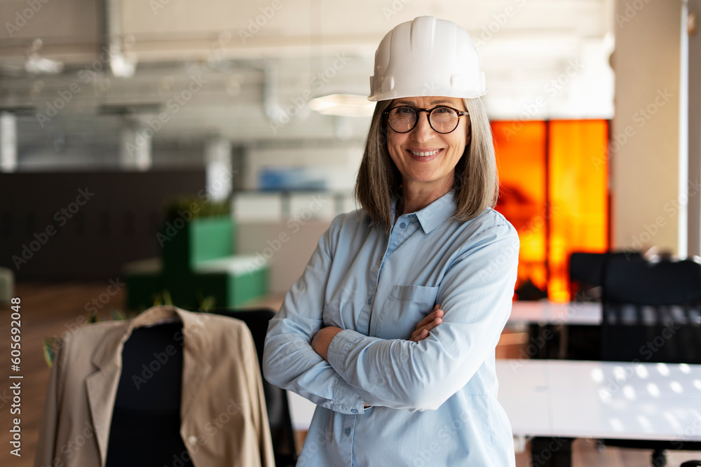 Portrait of confident smiling senior woman, architect, engineer holding arms crossed looking at camera standing in modern office. Successful business concept Portrait of confident smiling senior woman, architect, engineer holding arms crossed looking at camera standing in modern office. Successful business concept