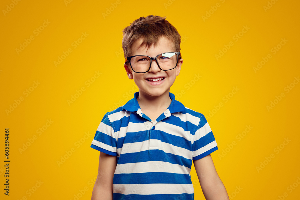 Cute positive kid schoolboy in blue striped polo shirt and glasses smiling and looking at camera while standing against yellow background