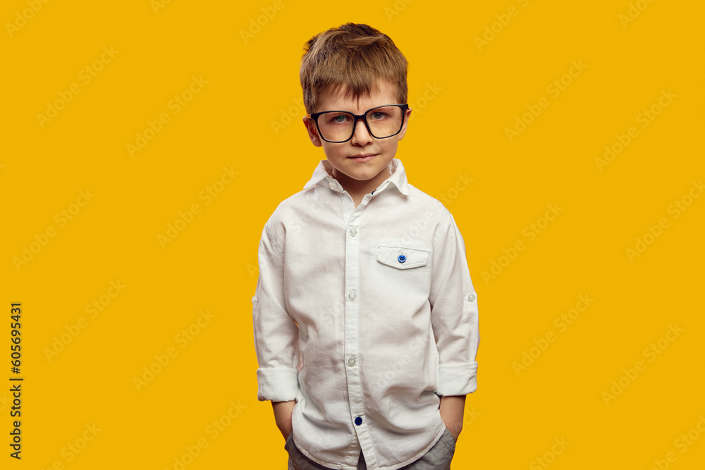 Fototapeta premium Serious little boy wearing white shirt and eyeglasses, keeping hands in pockets and looking mad at camera against yellow background