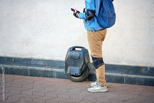 Boy ready to ride on monowheel. Man in protective gear stands near parked electric unicycle (EUC). Man wear protective knee pads, mono wheel parked against wall. Parked single wheel. Selective focus