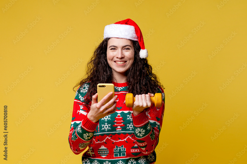 young girl in christmas clothes and santa hat trains for fitness and uses smartphone on yellow background young girl in christmas clothes and santa hat trains for fitness and uses smartphone on yellow background