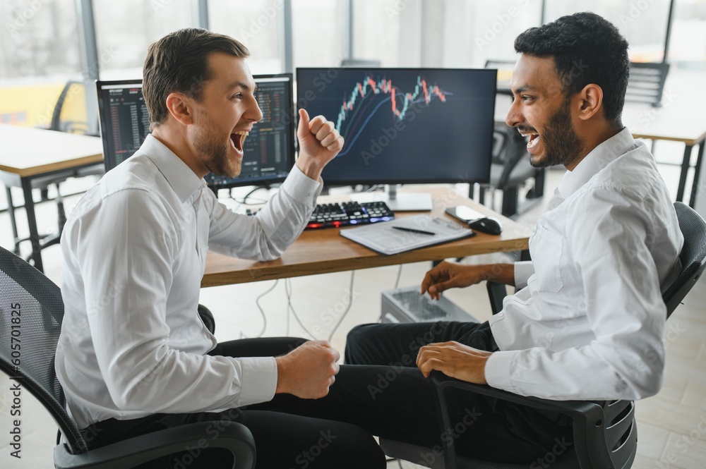 Fototapeta premium Two men traders sitting at desk at office together monitoring stocks data candle charts on screen analyzing price flow smiling cheerful having profit teamwork concept