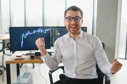 Man trader in formalwear sitting at desk in frot of monitors with charts and data at office browsing laptop checking documents analyzing stocks price changes concentrated. photo