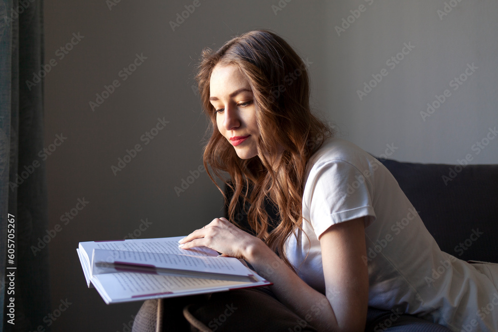 pensive woman in white t-shirt reads book at home and lies on the sofa, girl is focused on reading looks into the book pensive woman in white t-shirt reads book at home and lies on the sofa, girl is focused on reading looks into the book