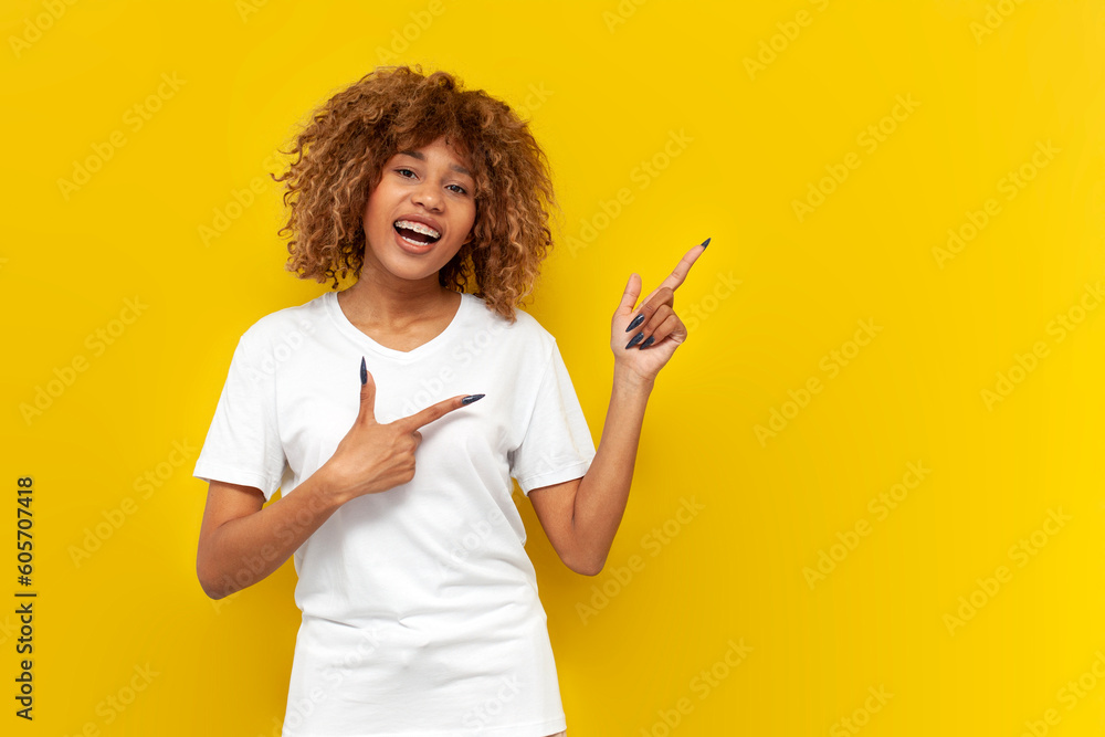 young curly american girl in white t-shirt and with braces points fingers to the side on yellow isolated background young curly american girl in white t-shirt and with braces points fingers to the side on yellow isolated background