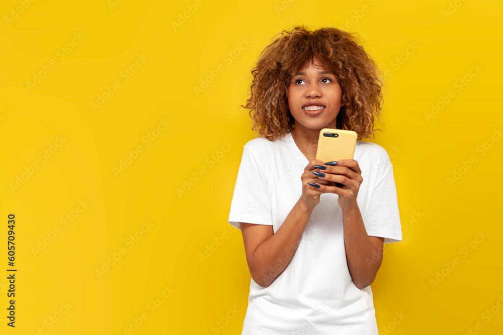 young curly american girl with braces holds smartphone and dreams on yellow isolated background young curly american girl with braces holds smartphone and dreams on yellow isolated background