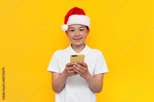 little asian boy in santa hat uses smartphone on yellow isolated background, korean child in new year's hat