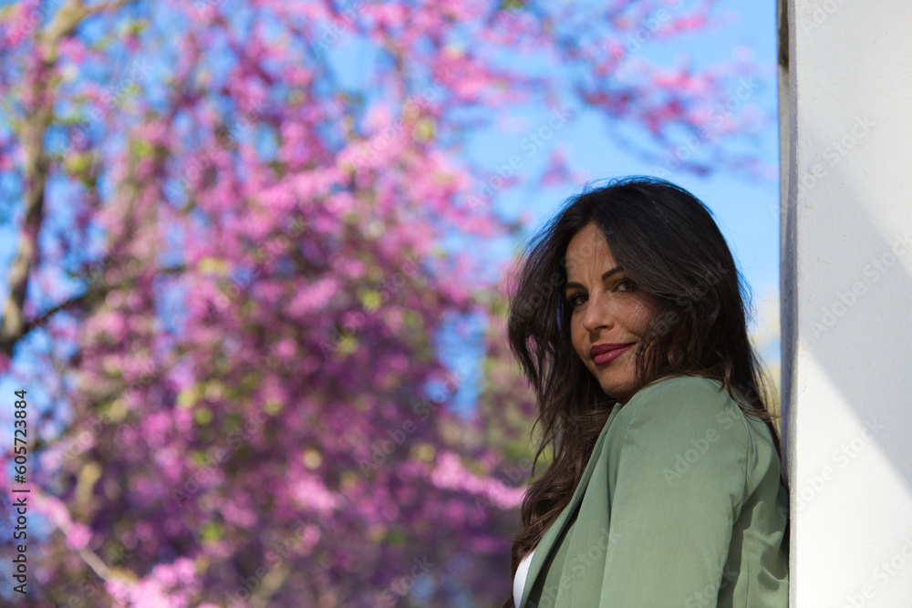 Portrait of beautiful young woman with long brown hair smiling and looking at the camera. In the background pink flowers and blue sky. Concept happiness and joy. Portrait of beautiful young woman with long brown hair smiling and looking at the camera. In the background pink flowers and blue sky. Concept happiness and joy.