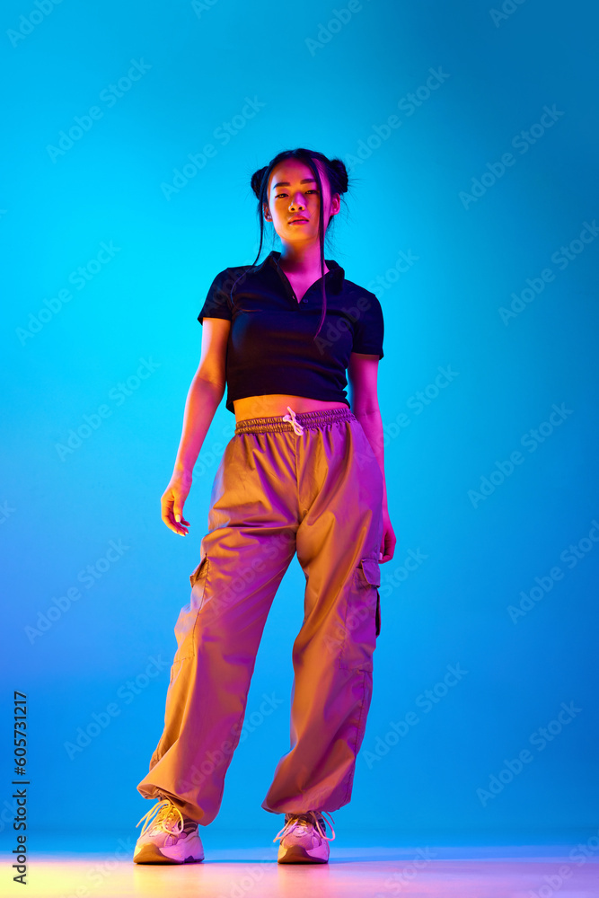 Full-length portrait of pretty korean girl posing in stylish casual clothes against blue studio background in neon light. Concept of emotions, facial expression, youth, lifestyle, inspiration, fashion