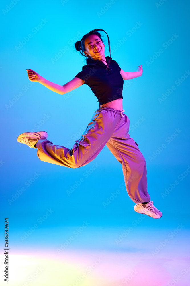 Full-length portrait of cheerful, happy, smiling korean girl jumping against blue studio background in neon light. Concept of emotions, facial expression, youth, lifestyle, inspiration, sales, ad