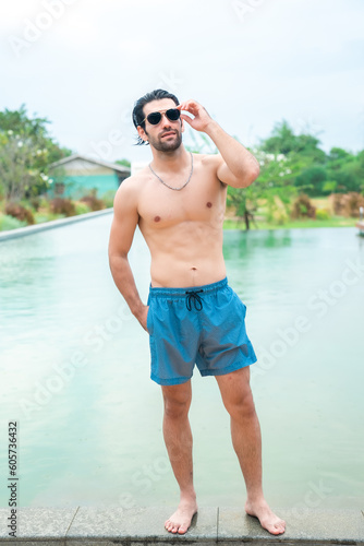 Sexy portrait of muscular handsome topless male model with perfect body in swimwear at swimming pool, attractive young man person with fit muscle posting relax lifestyle in summer vacation time