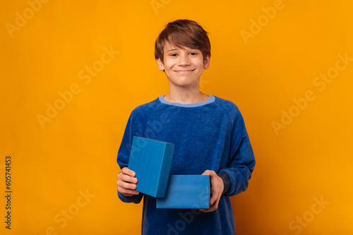 Cute smiling teen boy is holding a blue small gift box over yellow backdrop.