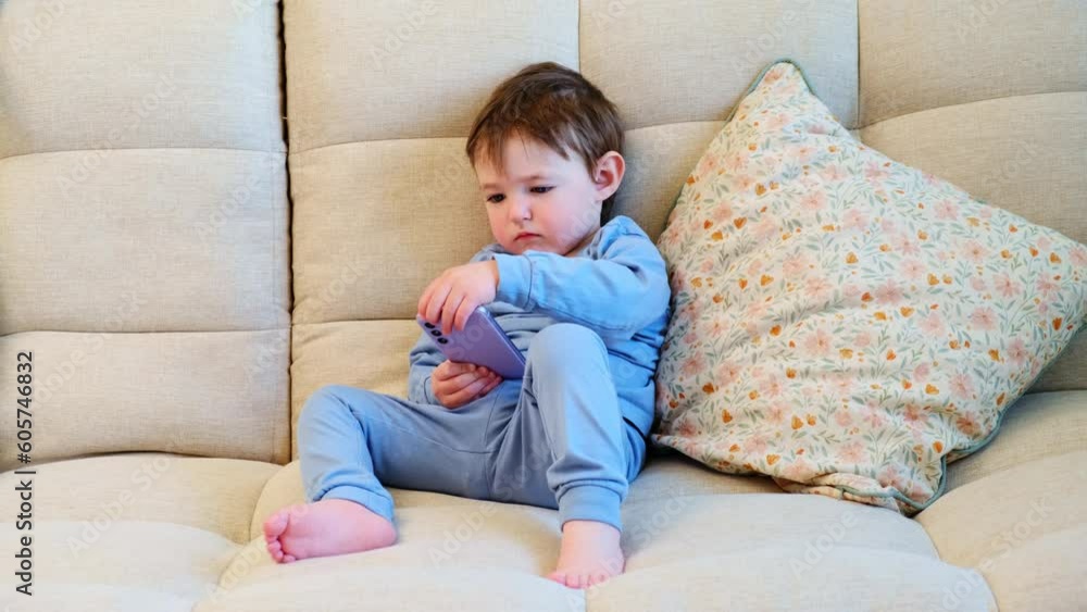 Toddler baby is tapping away on the mobile phone screen. The happy youngster is glued to the screen of their phone while seated comfortably on the couch. Kid aged about two years (one year ten months) Toddler baby is tapping away on the mobile phone screen. The happy youngster is glued to the screen of their phone while seated comfortably on the couch. Kid aged about two years (one year ten months)
