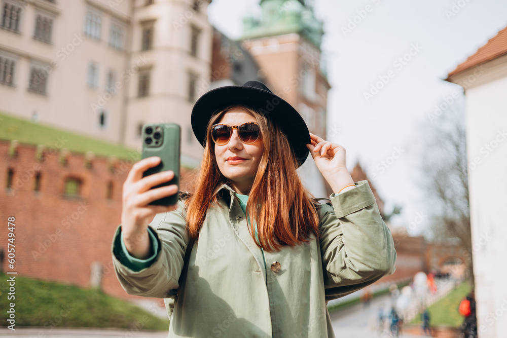 Fototapeta premium Attractive young female tourist is exploring new city. Redhead girl holding a smartphone in Krakow. Traveling. Happy optimistic girl walking in city and makes selfie poses against historic building