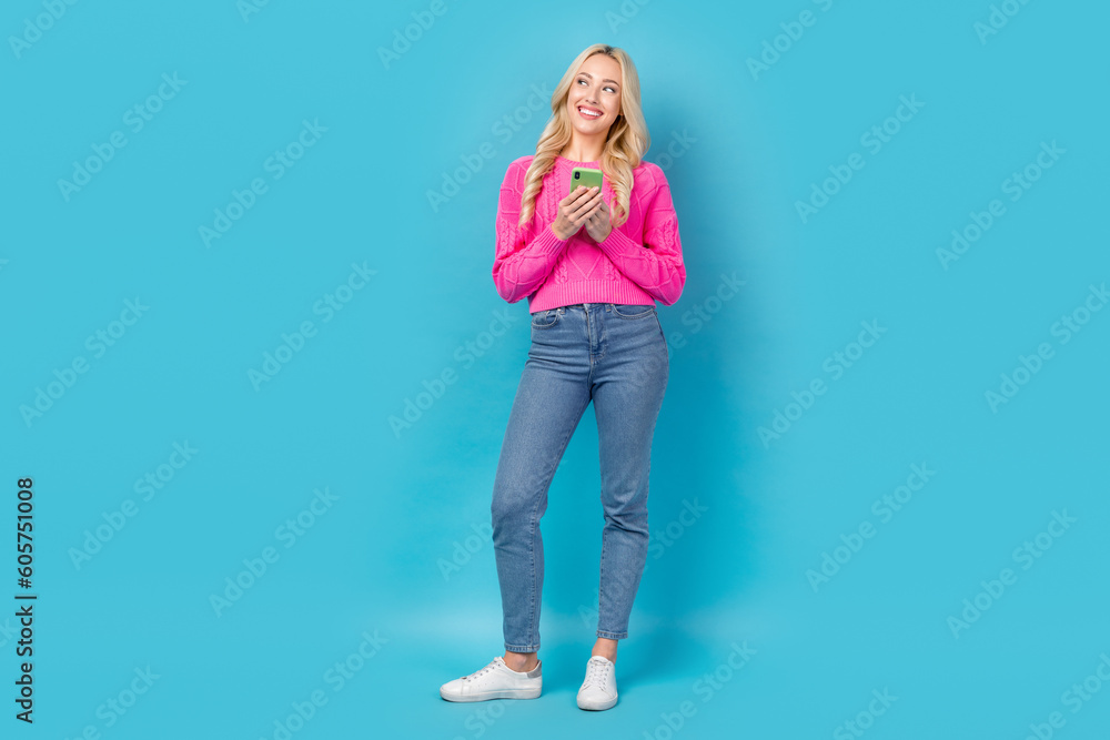 Full length photo of pretty person dressed knit sweater denim trousers hold smartphone look empty space isolated on blue color background Full length photo of pretty person dressed knit sweater denim trousers hold smartphone look empty space isolated on blue color background
