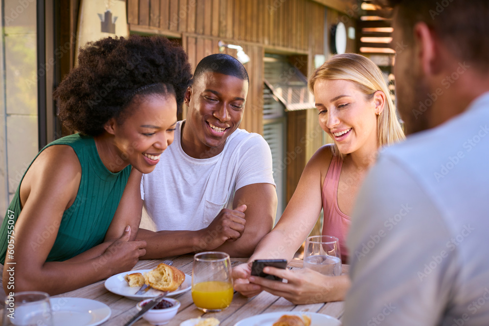 Group Of Smiling Multi-Cultural Friends Eating Breakfast Outdoors At Home Looking At Mobile Phone