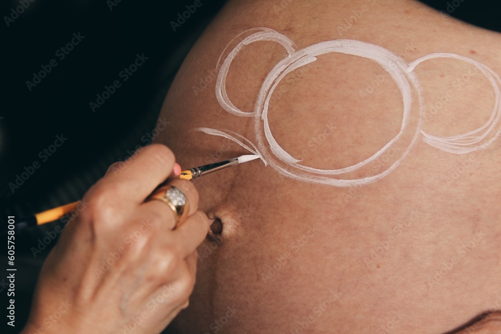 Female artist painting the belly of a pregnant young woman with body paints and brushes Female artist painting the belly of a pregnant young woman with body paints and brushes